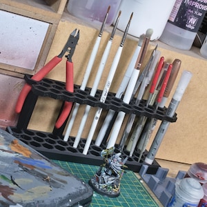 Könnte beinhalten: Ein schwarzer, sechseckiger Organizer hält verschiedene Kunstutensilien, darunter Pinsel mit weißen Griffen, Zangen mit roten Griffen und Stifte. Eine Miniaturfigur und eine Farbpalette sind ebenfalls zu sehen, was auf ein Bastel- oder Hobbythema hindeutet.