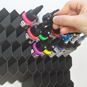 Op de afbeelding: Een zwarte, honingraatvormige organizer bevat verschillende kleurrijke inktflessen. De flessen hebben zwarte doppen en zijn gevuld met verschillende kleuren, waaronder roze, groen en blauw. Een hand reikt naar een van de flessen.