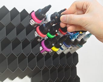 Op de afbeelding: Een zwarte, honingraatvormige organizer bevat verschillende kleurrijke inktflessen. De flessen hebben zwarte doppen en zijn gevuld met verschillende kleuren, waaronder roze, groen en blauw. Een hand reikt naar een van de flessen.