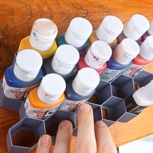 May include: A gray hexagonal organizer holds a collection of small paint bottles with white lids. The bottles contain various colors of paint, including blue, red, yellow, and green.