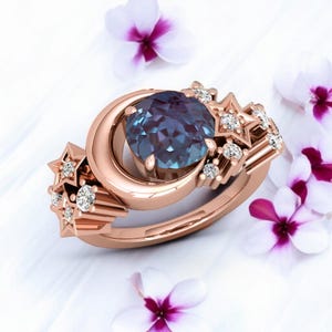 May include: Rose gold ring featuring a large, round, blue-green gemstone set within a crescent moon design. The ring is adorned with star-shaped accents and small, sparkling diamonds, creating a celestial theme.