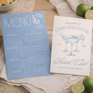 May include: Two printed menus on a wooden surface. One is light blue with a menu and food descriptions. The other is off-white with "Welcome to Cabo San Lucas" and "Social Club" text, plus cocktail illustrations.