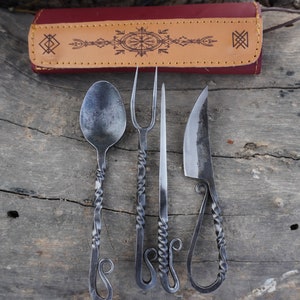 May include: A set of four hand-forged cutlery pieces, including a spoon, two forks, and a knife, with twisted handles and decorative loops. The cutlery is laid out on a wooden surface.