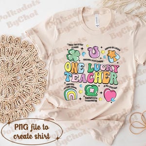 May include: A beige t-shirt featuring a colourful graphic design that reads "One Lucky Teacher". The graphic includes shamrocks, rainbows, stars, and hearts. The text "I teach the cutest clovers", "Teaching lucky charms", "Teaching future leaders", "Shenanigans coordinator", "Happy go lucky", and "In my lucky bra" are also included in the design.