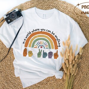 Peut inclure: T-shirt blanc avec un motif arc-en-ciel color&eacute; et le texte "in a world where you can be anything". Le motif comprend des gestes de la main et un c&oelig;ur. Un appareil photo vintage est pos&eacute; sur le t-shirt.