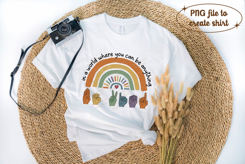 Peut inclure: T-shirt blanc avec un motif arc-en-ciel color&eacute; et le texte "in a world where you can be anything". Le motif comprend des gestes de la main et un c&oelig;ur. Un appareil photo vintage est pos&eacute; sur le t-shirt.