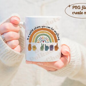 Peut inclure: Mug en c&eacute;ramique blanche avec un motif arc-en-ciel et le texte "in a world where you can be anything". Le motif comprend des signes de la main et un c&oelig;ur. La tasse est tenue par une personne.
