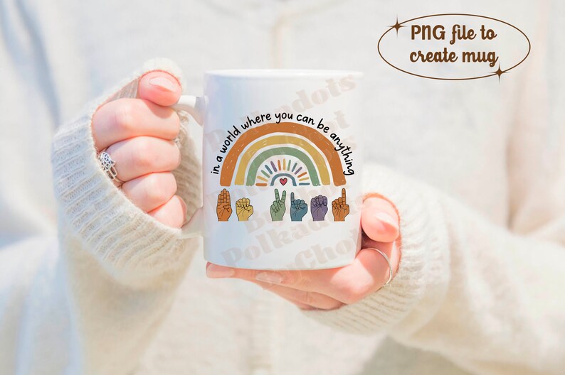 Peut inclure: Mug en c&eacute;ramique blanche avec un motif arc-en-ciel et le texte "in a world where you can be anything". Le motif comprend des signes de la main et un c&oelig;ur. La tasse est tenue par une personne.