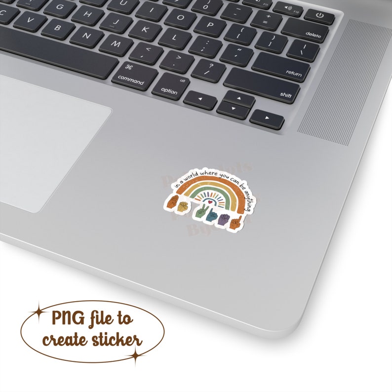 Peut inclure: Un autocollant avec un motif arc-en-ciel et le texte "in a world where you can be anything". L'autocollant, avec des symboles de mains, est sur un ordinateur portable. Le fichier est un PNG pour cr&eacute;er un autocollant.
