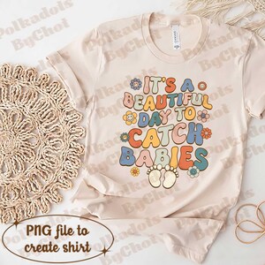 May include: Cream-colored t-shirt with the text "It's a beautiful day to catch babies" in colorful, retro-style lettering. The design includes flower and footprint graphics, perfect for a baby shower or new parent.