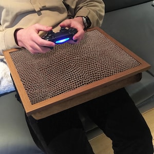 Puede incluir: Un escritorio portátil de madera con una superficie texturizada marrón, diseñado para jugar o trabajar. El escritorio tiene un marco marrón claro y una base acolchada. Una persona sostiene un mando de juego negro con una luz azul.