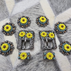 May include: A set of nine silver-toned metal belt buckles with sunflower designs. The buckles are decorated with blue and yellow rhinestones.