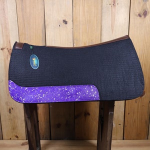 May include: A black saddle pad with a purple and white speckled trim. The saddle pad has a brown leather binding and a small green star on the top left corner.