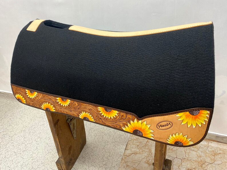 May include: Black saddle pad with a tan leather border and sunflower design. The border is tooled with a floral pattern. The saddle pad has the word "Mousy" printed on it.