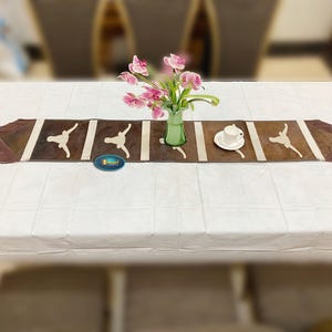 May include: A brown table runner with white stripes and a fringed edge, decorated with stylized figures. A vase of pink flowers and a teacup and saucer sit on the runner, which is placed on a white tablecloth.