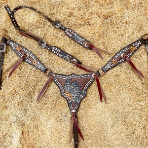 Op de afbeelding: Een bruin en wit leren paardentuigset met decoratieve zilverkleurige accenten. De set bevat een hoofdstel, borsttuig en teugels, met bloemen en geometrische ontwerpen. Het borsttuig heeft een centraal veermotief.