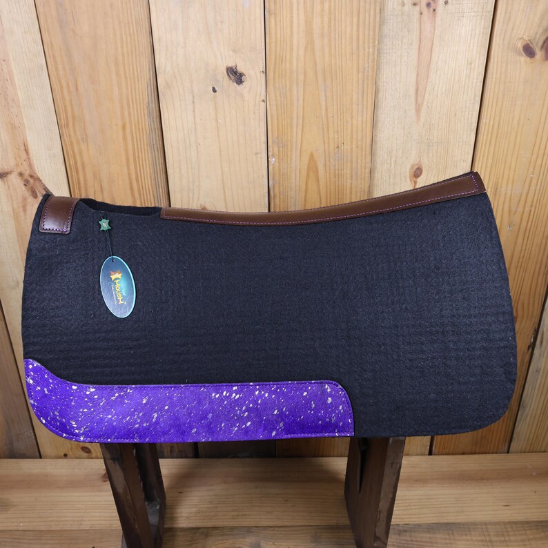 May include: A black saddle pad with a purple and white speckled trim. The saddle pad has a brown leather binding and a green star on the top left corner. The saddle pad is on a wooden surface.