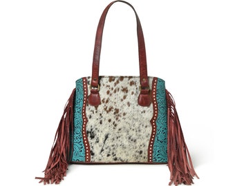 Puede incluir: Un bolso tote de piel de vaca con detalles turquesa y asas de cuero marrón. El bolso presenta un estampado moteado y flecos decorativos en los laterales. Las asas están unidas con anillos color latón.