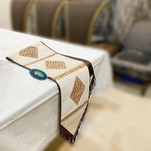 Puede incluir: Un camino de mesa blanco con ribete marrón y un diseño de estampado de leopardo. El camino de mesa tiene flecos en los extremos y está hecho de una tela suave y texturizada.
