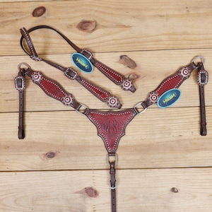 Peut inclure: Un ensemble de harnachement de cheval de style western avec une poitrail en cuir rouge et marron, un frontal et des rênes. L'ensemble est orné d'éléments argentés et d'ornements floraux. Le poitrail présente un motif estampé et l'étiquette indique "Housh".