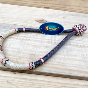 Op de afbeelding: Een zwart en wit gevlochten leren touw met rode en witte accenten. Het touw heeft de vorm van een cirkel en heeft een decoratief knoopje aan het einde. Het touw ligt op een houten oppervlak.