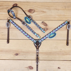 Op de afbeelding: Een paardentuigset met een gevlochten borsttuig, frontriem en teugels. De set is in tinten paars en turquoise, met zilverkleurige hardware en decoratieve accenten. Het leer is bruin, en de set bevat labels met de merknaam.