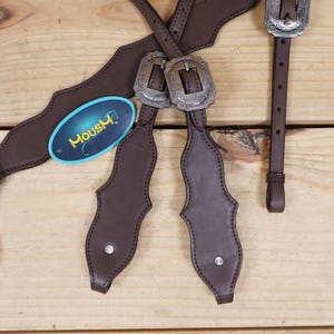 Puede incluir: Primer plano de un equipo de caballo de cuero marr&oacute;n con hebillas plateadas y detalles decorativos. El equipo incluye correas y una etiqueta con la palabra "Moushy". El fondo es una superficie de madera clara.