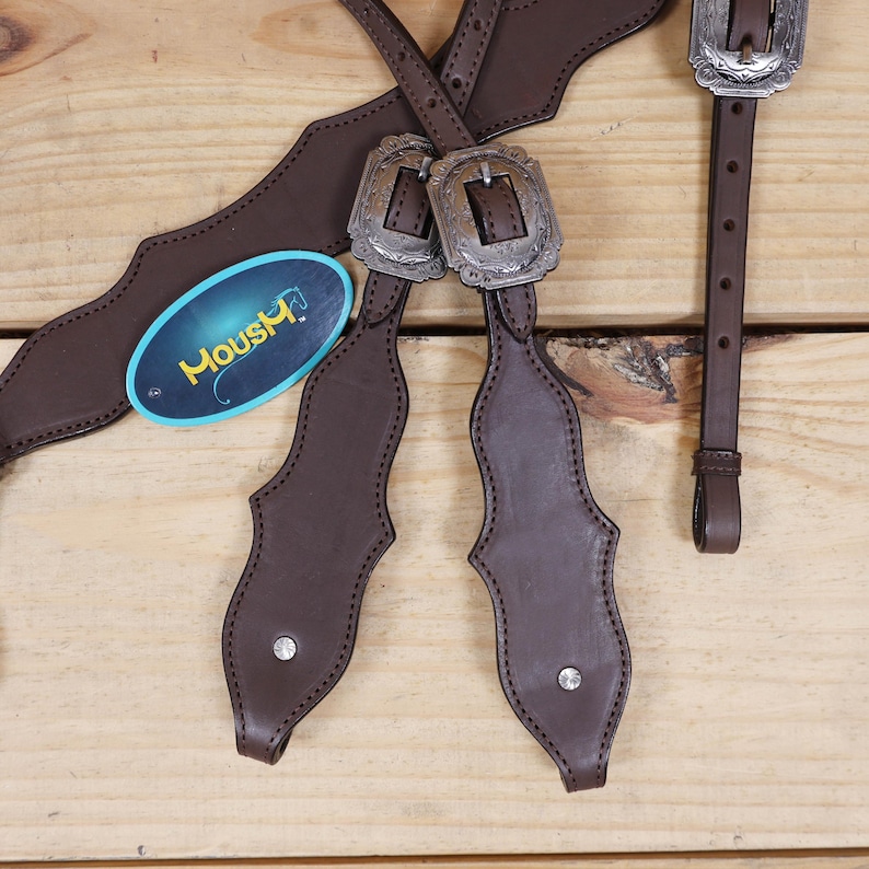 Puede incluir: Primer plano de un equipo de caballo de cuero marr&oacute;n con hebillas plateadas y detalles decorativos. El equipo incluye correas y una etiqueta con la palabra "Moushy". El fondo es una superficie de madera clara.