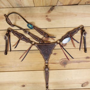 Könnte beinhalten: Ein braunes Leder-Pferdegeschirr-Set mit aufwendigen Blumen- und Sternmuster-Designs. Das Set umfasst einen Kopfhalfter, einen Brustgeschirr und Zügel, alle mit silberfarbener Hardware und dekorativen Akzenten. Das Leder ist dunkelbraun.