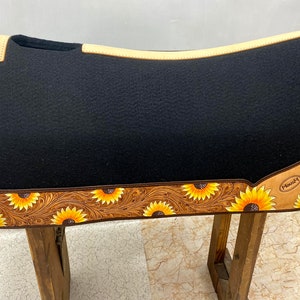 May include: Black saddle pad with a brown leather border and sunflower design. The border is tooled with a floral pattern. The saddle pad is made by the brand "Maxam".
