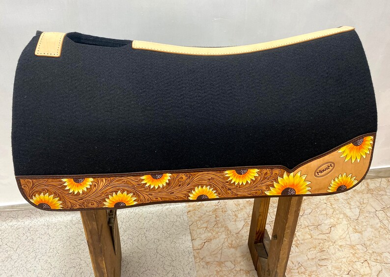 May include: Black saddle pad with a brown leather border and sunflower design. The border is tooled with a floral pattern. The saddle pad is made by the brand "Maxam".