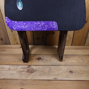 May include: A black and purple saddle pad with a white splatter design. The saddle pad is on a wooden stand.