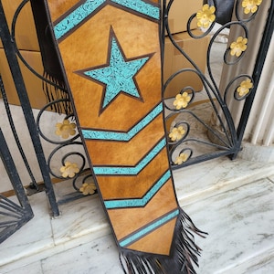 May include: A brown leather table runner with a turquoise star and chevron pattern. The runner has black fringe on the ends.