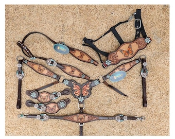 Puede incluir: Un conjunto de guarnicionería para caballos en cuero marrón con patrones de mariposas en relieve, conchos florales de color turquesa y herrajes plateados. El set incluye una cabezada, un pretal y correas para espuelas con motivos de mariposas.