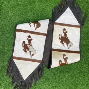 以下が含まれることがあります： フリンジの縁取りが施された白と茶色の牛革のテーブルランナー。ランナーには、馬に乗ったカウボーイが繰り返し描かれています。