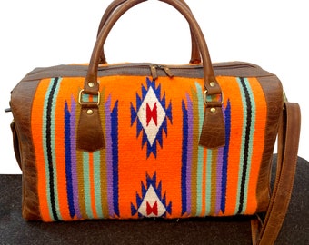 Op de afbeelding: Een bruine leren reistas met een geweven geometrisch patroon in oranje, blauw en paars. De tas heeft twee bovenste handvatten en een lange schouderband.