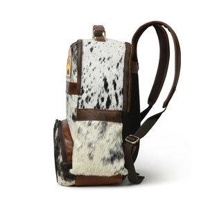 May include: A brown leather backpack with a white and black cowhide pattern. The backpack has a large main compartment, a front pocket, and two adjustable shoulder straps.