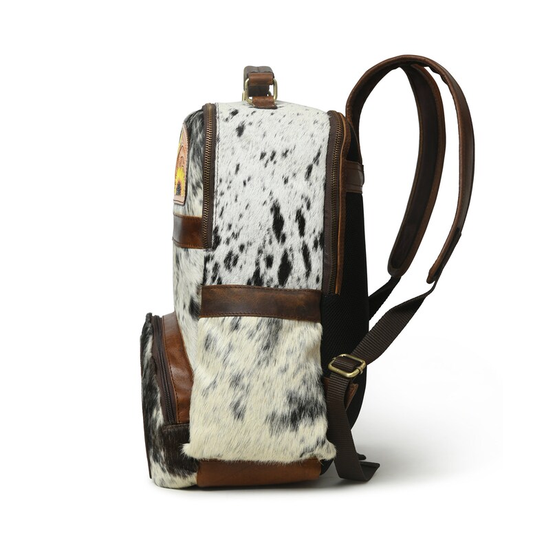 May include: A brown leather backpack with a white and black cowhide pattern. The backpack has a large main compartment, a front pocket, and two adjustable shoulder straps.