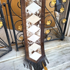 May include: A brown and white cowhide table runner with fringe. The runner is made of leather and has a diamond pattern.