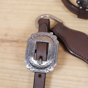 Puede incluir: Primer plano de una brida de caballo de cuero marr&oacute;n con una hebilla decorativa plateada. La hebilla tiene un dise&ntilde;o floral ornamentado. La brida est&aacute; sobre un fondo de madera clara.