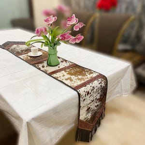 Puede incluir: Un camino de mesa blanco con un patrón de piel de vaca marrón y blanco y ribete de flecos. El camino de mesa está sobre un mantel blanco con un jarrón de flores rosas en el centro.