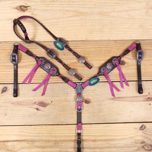 Könnte beinhalten: Ein braunes und rosa Pferdezubehör-Set mit silberfarbenen Akzenten. Das Set enthält einen Kopfhalfter, einen Brustgeschirr und Zügel, alle mit dekorativen silbernen Conchos und rosa Akzenten. Das Zubehör wird auf einer Holzoberfläche präsentiert.