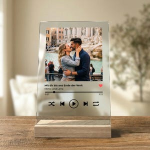Könnte beinhalten: Eine transparente Acryl-Fotodisplay mit Holzsockel. Das Display zeigt ein Foto eines sich küssenden Paares vor einem Brunnen. Der Text auf dem Display lautet "Mit dir bis ans Ende der Welt Mirko und Lena".