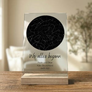 Könnte beinhalten: Transparente Acrylplatte mit Sternenkarte in Schwarz-Weiß, montiert auf einem hellen Holzsockel. Der Text "Wo alles begann" steht über Namen, Ort und Datum.