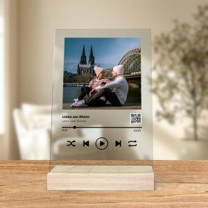 Könnte beinhalten: Ein gläserner Musikplayer mit einem Foto eines Paares, das auf einer Brücke vor einer Kathedrale sitzt. Der Musikplayer hat einen schwarzen Hintergrund mit weißem Text, der "Liebe am Rhein" und "Lena und Marlon" lautet. Der Musikplayer hat außerdem einen Play-Button, einen Pause-Button, einen Rückwärts-Button, einen Vorwärts-Button und einen Repeat-Button.