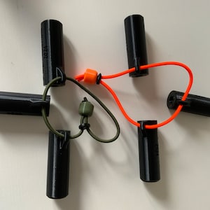 Könnte beinhalten: Fünf schwarze zylindrische Objekte mit einer kleinen Schlaufe oben sind durch zwei Schnüre verbunden. Eine Schnur ist orange und die andere grün. An der orangen Schnur befindet sich eine kleine orange Perle und an der grünen Schnur eine kleine grüne Perle.