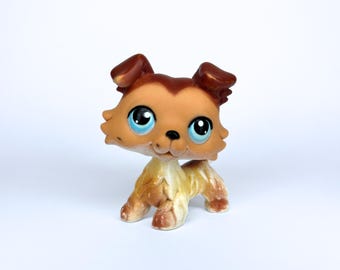May include: A tan and brown dog figurine with large blue eyes. The toy dog has a light tan body and a brown head and ears. The figurine is standing on all fours and has a friendly expression.