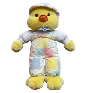 May include: A plush yellow chick wearing a light blue hat and bow tie, and patchwork overalls. The chick has a round, fluffy body, orange beak, and black eyes. The overalls are made of pastel-colored fabric squares.