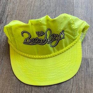 May include: A neon yellow nylon cap featuring a braided cord trim across the base of the crown. The front of the cap displays the text The Beach Boys embroidered in black thread with a light grey outline, centered above the curved brim.