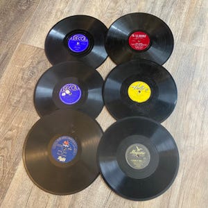 May include: Six vintage vinyl records are arranged on a wooden surface. The records are black with colorful labels, including blue, red, and yellow. The labels display text such as "Decca", "Waldorf", and "Majestic".
